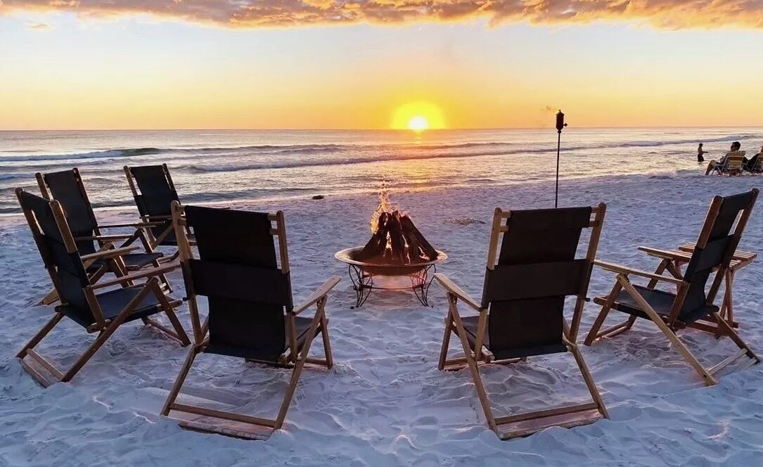 A beach scene with four wooden chairs arranged around a central fire pit, overlooking the ocean at sunset.