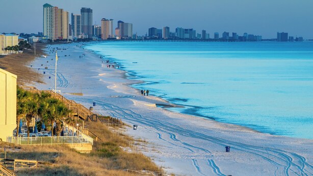 Ein Strand mit einer Stadtansicht im Hintergrund, einer Promenade mit Palmen und einem sandigen Strand mit Fußabdrücken.