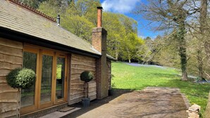 A log cabin with a chimney, wooden siding, and a small garden with potted plants. - Sanctuary in the Surrey Hills, Secluded Woodland Cottage, Walking Cycling Nature (Ranmore Common)