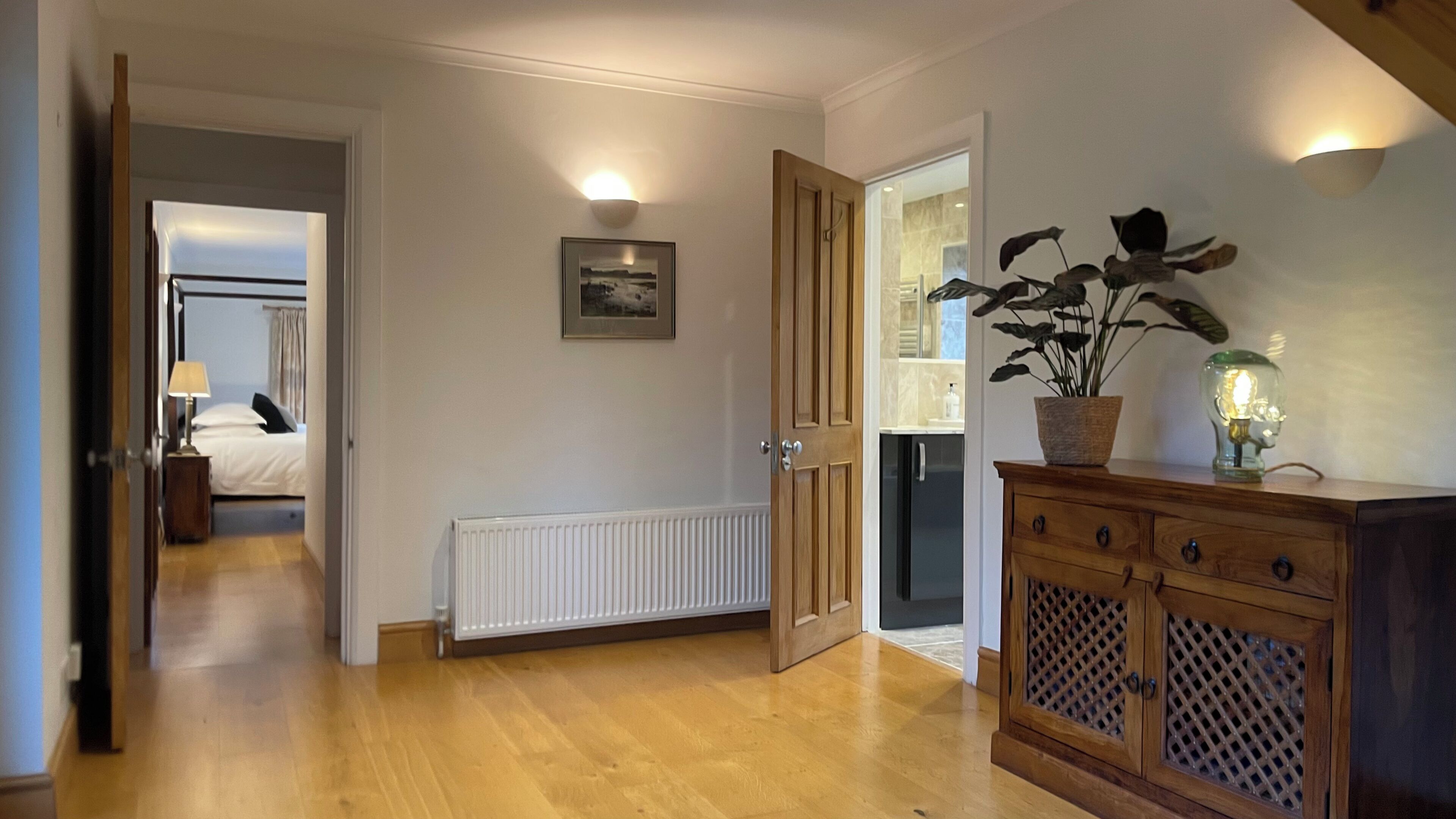 A hallway with a wooden floor, a radiator, a door leading to a room, a cabinet with a plant, and a wall-mounted light fixture.