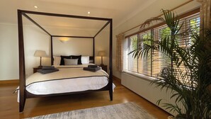 A four-poster bed with a white and black bedspread, two bedside lamps, a large potted plant, and a window with blinds. - Sanctuary in the Surrey Hills, Secluded Woodland Cottage, Walking Cycling Nature (Ranmore Common)