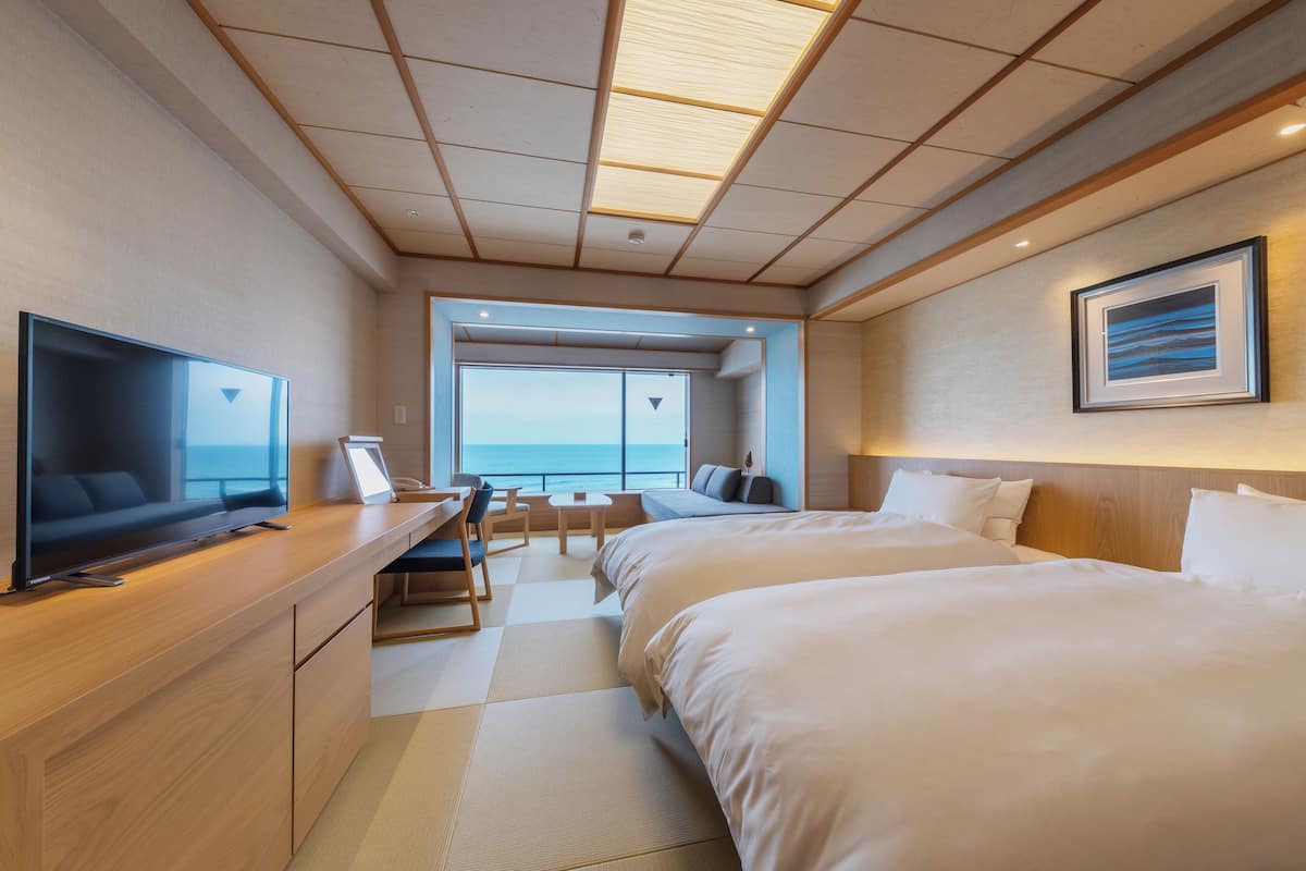 Une chambre d’hôtel moderne dotée d’un grand lit, d’un bureau avec un téléviseur à écran plat, d’un coin salon offrant une vue sur la mer et d’un tableau encadré au mur.