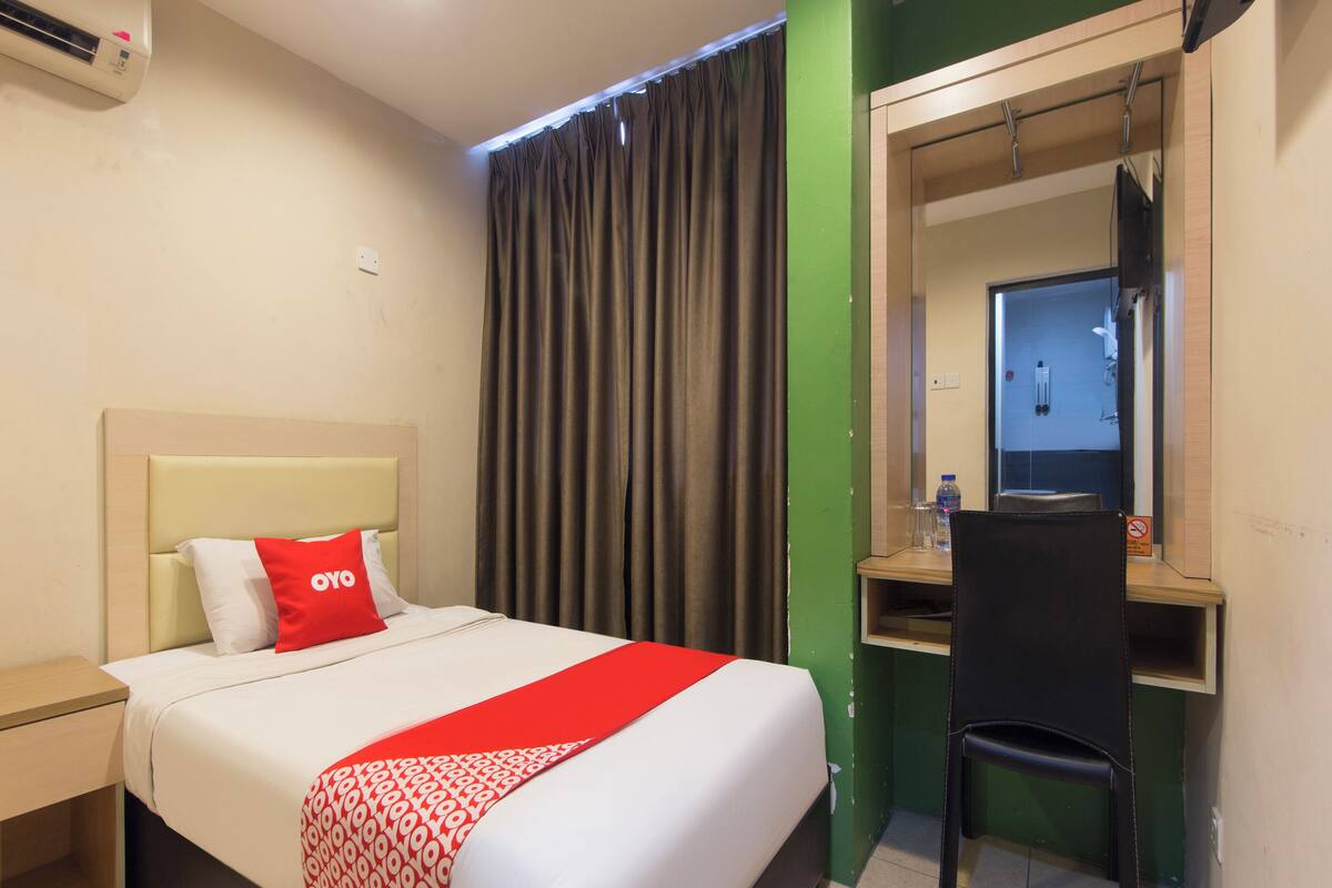 A hotel room with a bed, a desk, a chair, a mirror, and a bathroom visible through a glass door.