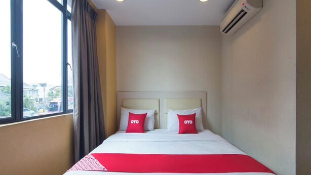 A hotel room with a bed, two pillows with the 'OYO' logo, a red and white bedspread, a window with curtains, and an air conditioning unit on the wall. - Hotel 916 (Batu Caves)