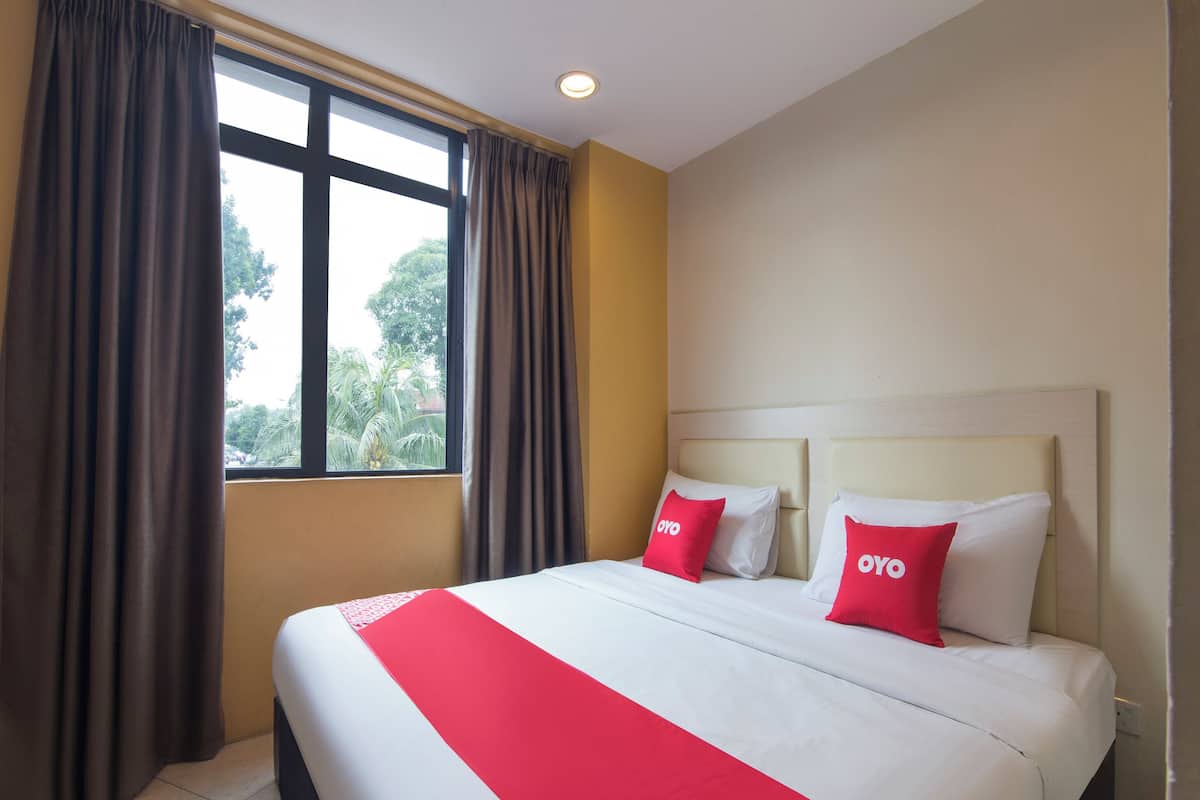 A hotel room with a large bed, two pillows with the brand 'OYO', a window with curtains, and a view of trees outside.