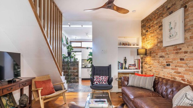 Un salon moderne avec un mur en briques, un escalier en bois, un canapé en cuir, une table basse en bois et un téléviseur.