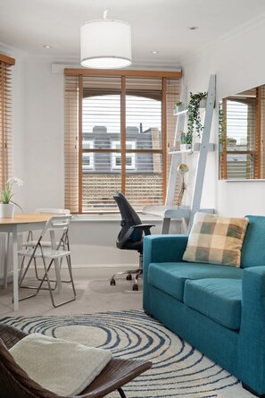 A modern living room with a blue sofa, a wooden dining table, and a white chair. There is a large window with blinds, a white pendant light, and a ladder leaning against the wall. - Stylish Comfortable 2BR Easy City Links (London)