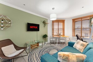 Ein modernes Wohnzimmer mit einer Couch, einem Sessel, einem Esstisch mit Stühlen, einem an der Wand montierten Fernseher und einem Fenster mit Jalousien.