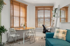 Ein modernes Wohnzimmer mit einem türkisfarbenen Sofa, weißen Stühlen, einem Esstisch und großen Fenstern mit Holzjalousien.