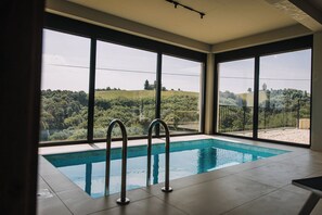 Une piscine dotée d’échelles en métal, de grandes fenêtres et offrant une vue sur un paysage verdoyant.