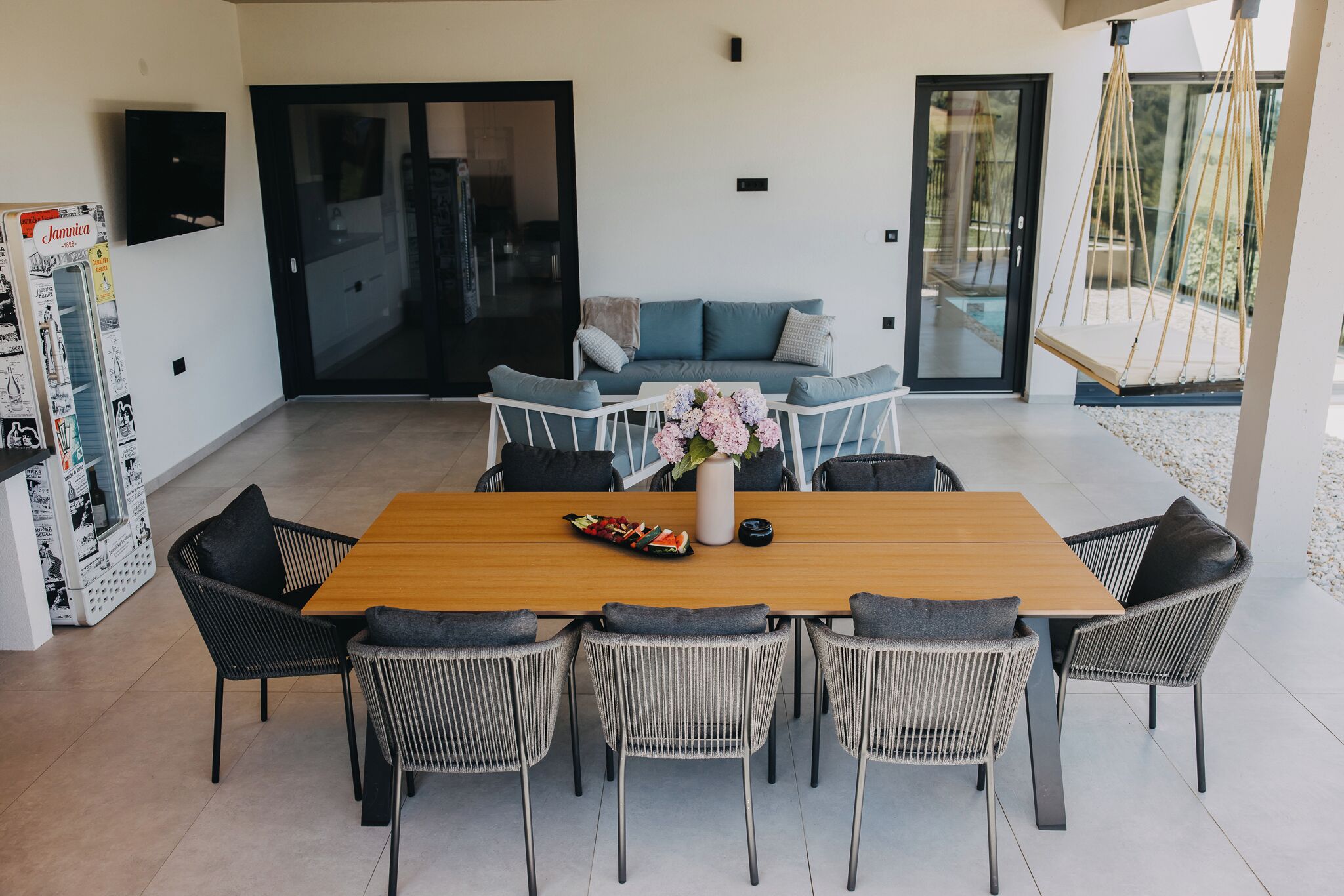 A modern dining area with a wooden table and wicker chairs, a sofa with cushions, and a hanging swing chair.