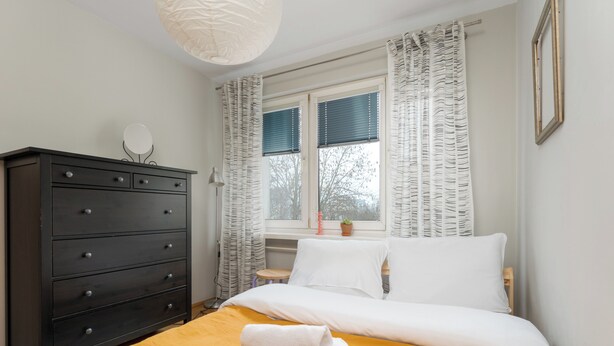 Un lit bien fait, recouvert d’une couvre-lit jaune, une commode en bois foncé avec des tiroirs, un miroir rond et une fenêtre avec des rideaux.