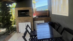 Patio esterno con tavolo e sedie in legno, barbecue a incasso e vista sul verde circostante.