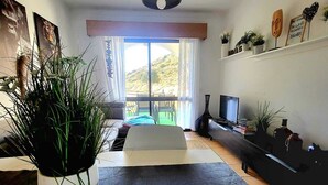 A modern living room with a large window, a potted plant, a white table, and a TV. - CHABELLA! Charm Beach Lofts Apartments - Garden & Sea View - CARVOEIRO - ALGARVE (Carvoeiro)