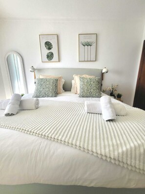 Une chambre à coucher avec un grand lit, deux oreillers, une tête de lit, un miroir et deux reproductions botaniques encadrées accrochées au mur.