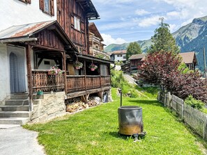 Una casa de madera tradicional con balcón, una parrilla pequeña al aire libre y montañas al fondo.
