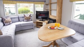 A modern living room with a grey sofa, a wooden coffee table with fruit and juice, a TV, and a view of greenery outside. - The Lake House - Quiet, Dog Friendly, By the Lake, Free Parking (Bowness-on-Windermere)