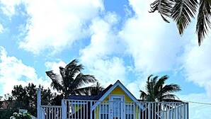 A two-story building with a balcony, surrounded by palm trees and a gravel driveway. - Cottage By The Sea ~Wonderful Family Friendly 2-bedroom with AC in East End (East End)
