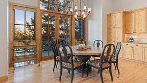 A dining area with a round table, chairs, a chandelier, wooden cabinets, and a view through large windows. - Abode at Elkhorn Village, hot tub, steps to skiing (Sun Valley)
