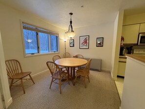 Ein Essbereich mit einem runden Tisch und sechs Stühlen, einem Fenster mit Jalousien, einer Stehlampe und einer im Hintergrund sichtbaren Küche.