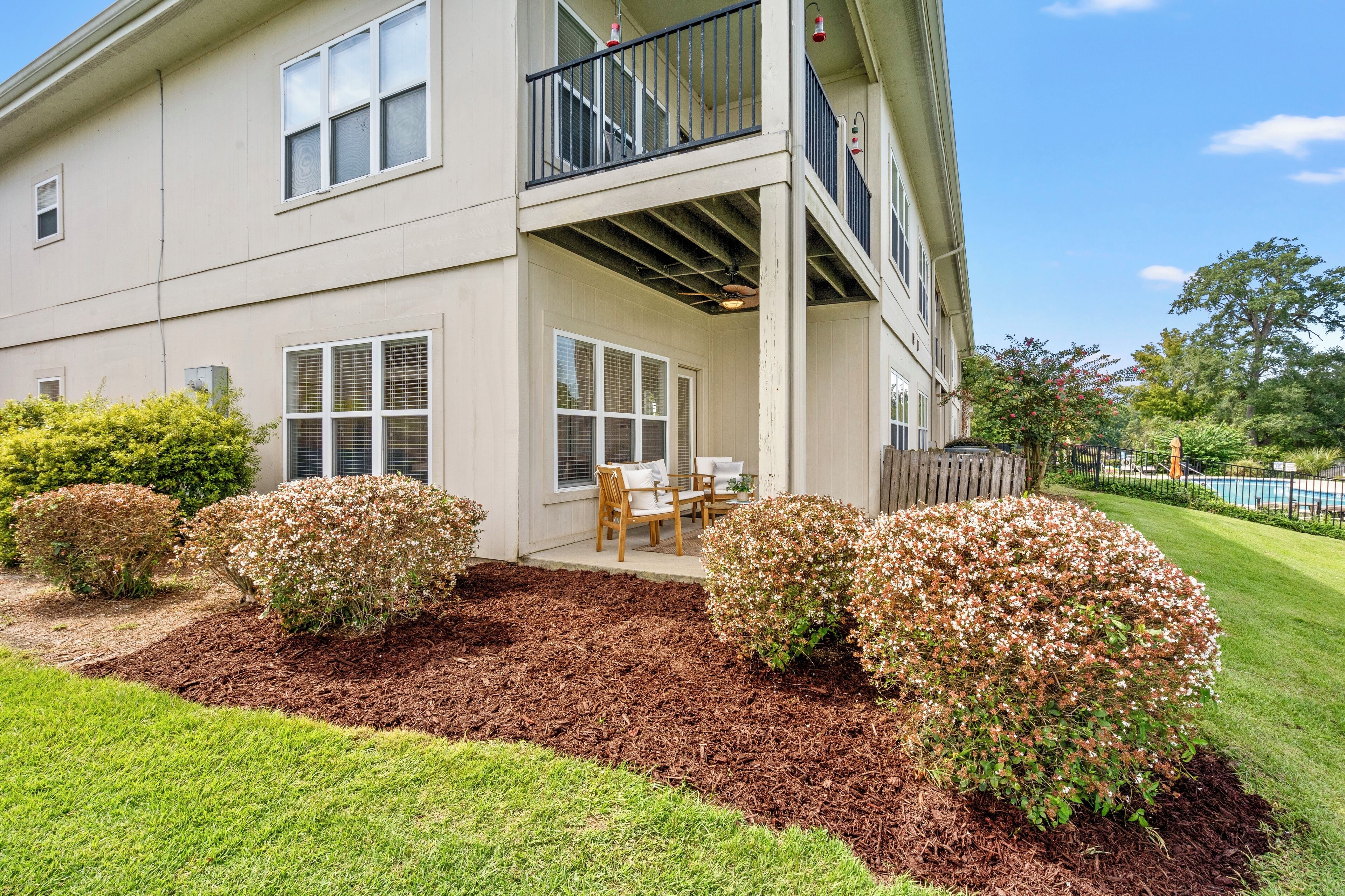 A two-story building with a balcony, a covered patio area with outdoor furniture, and a well-maintained garden with mulch and shrubs.