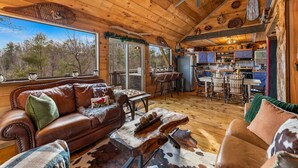A cozy cabin interior with a leather sofa, wooden dining table, and a kitchen area. - Bigfoot's Hideout - Treehouse, Arcade, Hot Tub (Blue Ridge)