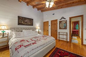 A bedroom with a large bed, wooden ceiling beams, a ceiling fan, a wall-mounted painting, and a wooden door. - Riverbend Lodge - Riverfront | Screened Area w Outdoor Fireplace | Firepit (Ellijay)