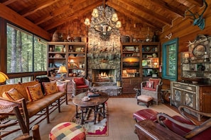 A cozy cabin interior with a stone fireplace, wooden furniture, and a large window overlooking a forest. - Riverbend Lodge - Riverfront | Screened Area w Outdoor Fireplace | Firepit (Ellijay)