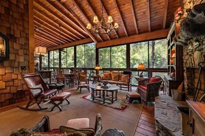 A spacious room with wooden flooring, a fireplace, and large windows offering a view of the forest. - Riverbend Lodge - Riverfront | Screened Area w Outdoor Fireplace | Firepit (Ellijay)