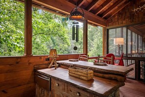 A rustic wooden interior with a large window overlooking a forest, a wooden table with chairs, and a hanging light fixture. - Riverbend Lodge - Riverfront | Screened Area w Outdoor Fireplace | Firepit (Ellijay)