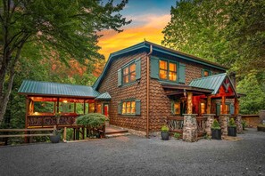 A rustic lodge with a stone facade, wooden deck, and a covered porch. - Riverbend Lodge - Riverfront | Screened Area w Outdoor Fireplace | Firepit (Ellijay)