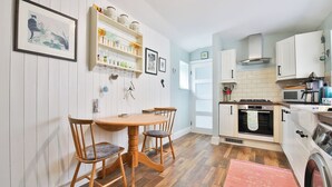 A kitchen with white cabinets, a wooden dining table with chairs, and a hanging plant shelf. - Bright Maisonette Near Beach and University (Swansea)