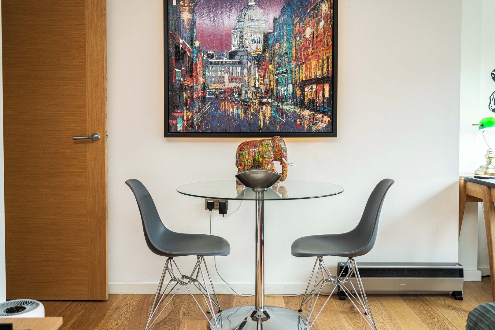 Een moderne kamer met een glazen tafel, twee zwarte stoelen, een kleurrijk olifantbeeld en een stadsgezicht schilderij in een lijst aan de muur.