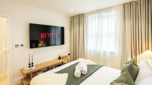 Quarto de hotel com cama, televisão na parede exibindo 'NETFLIX', um banco de madeira e uma janela com cortinas.