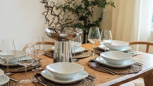 Mesa de jantar posta com pratos, tigelas e copos de vinho brancos, rodeada por jogos americanos trançados e talheres.