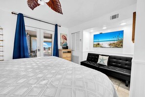 A bedroom with a large bed, a black leather sofa, a painting of a beach, and a view of the sea through the window. - Sanddollar 19B* Directly On The Beach! Amazing Views*Private Beach* (Miramar Beach)
