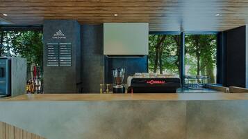 Un interno di caffetteria moderna con una bacheca dei prezzi, una macchina da caffè espresso La Cimbali e vista sugli alberi attraverso ampie finestre.