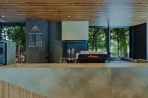 Interior moderno de una cafetería con un menú, una máquina de café espresso La Cimbali y vistas a árboles a través de grandes ventanales.