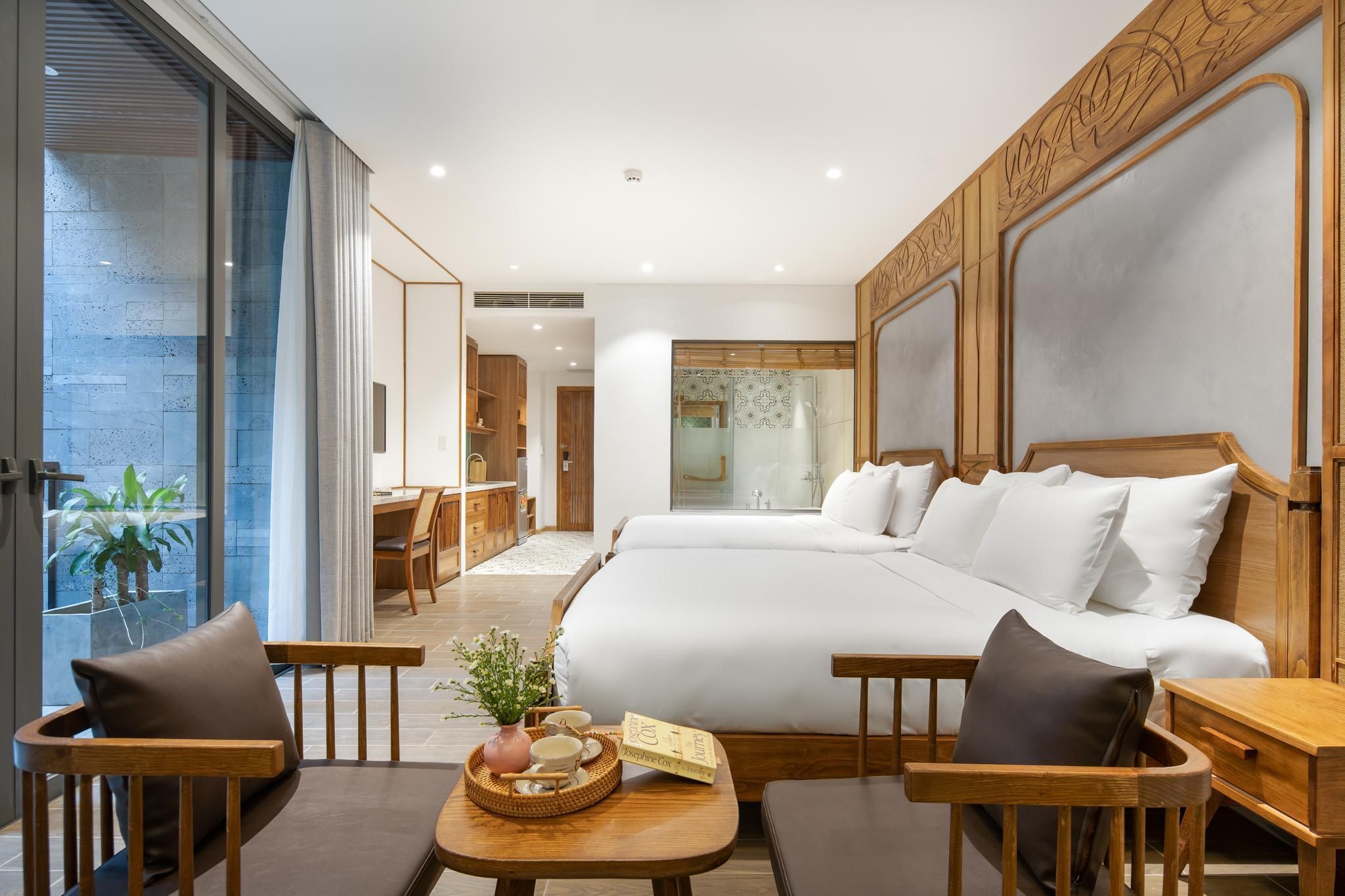 Una habitación de hotel moderna con una cama grande, muebles de madera, una mesita con un florero y un libro, y un balcón con plantas.