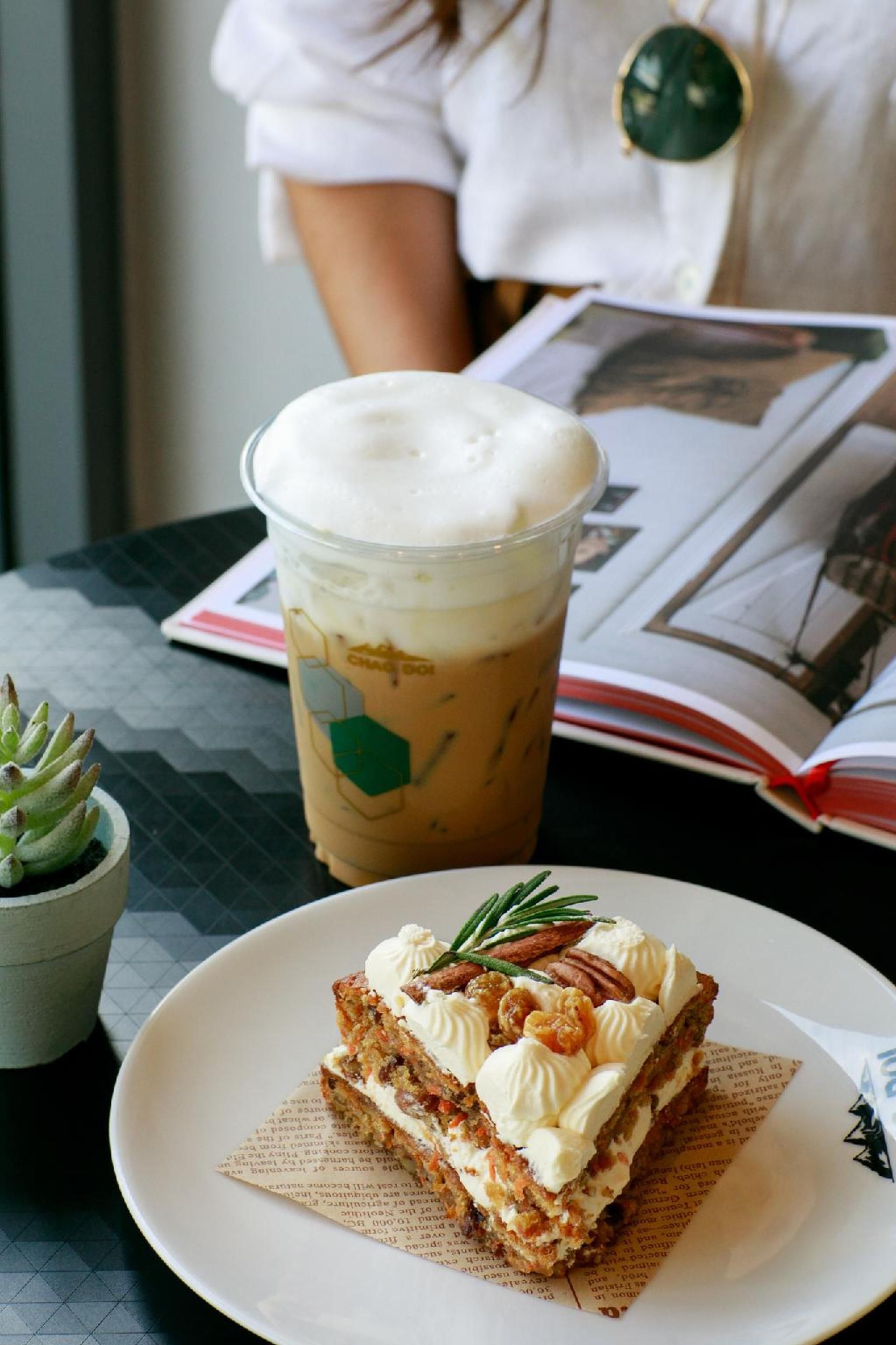 A slice of carrot cake with cream and nuts on a plate, a cup of iced coffee with whipped cream, and a small succulent plant on a table.