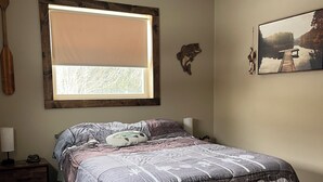 Un dormitorio con cama, ventana con persianas, ventilador de techo, un adorno de pez montado en la pared y un cuadro enmarcado.
