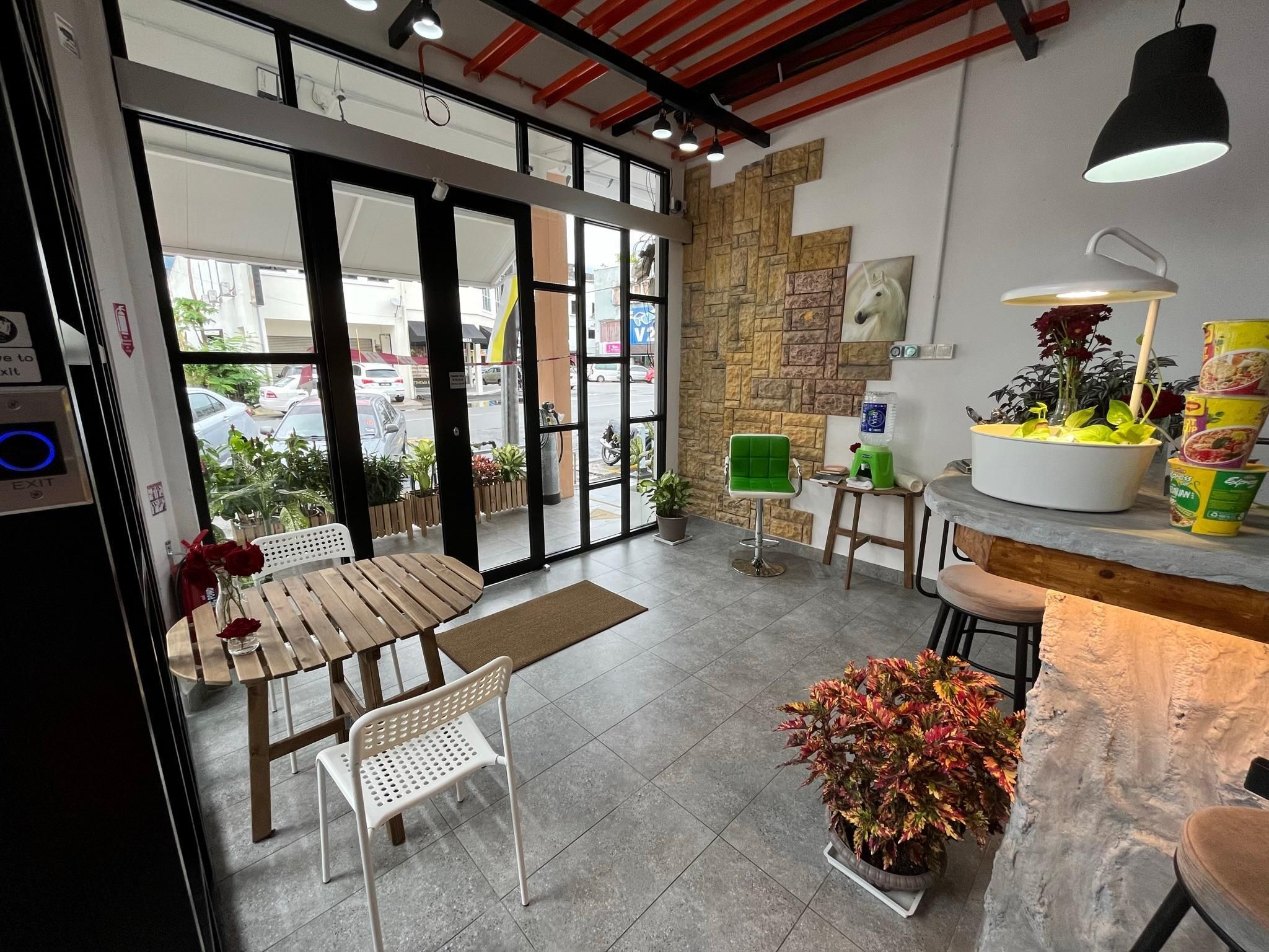 A cafe with a wooden table and chairs, a green chair, a potted plant, and a counter with a water dispenser and food containers.