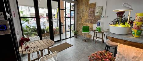 A cafe with a wooden table and chairs, a green chair, a potted plant, and a counter with a water dispenser and food containers.