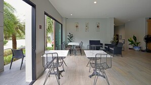 A modern indoor space with a dining area featuring metal chairs and white tables, a seating area with sofas, and a large potted plant. - Raweewan Residence (Bangkok)
