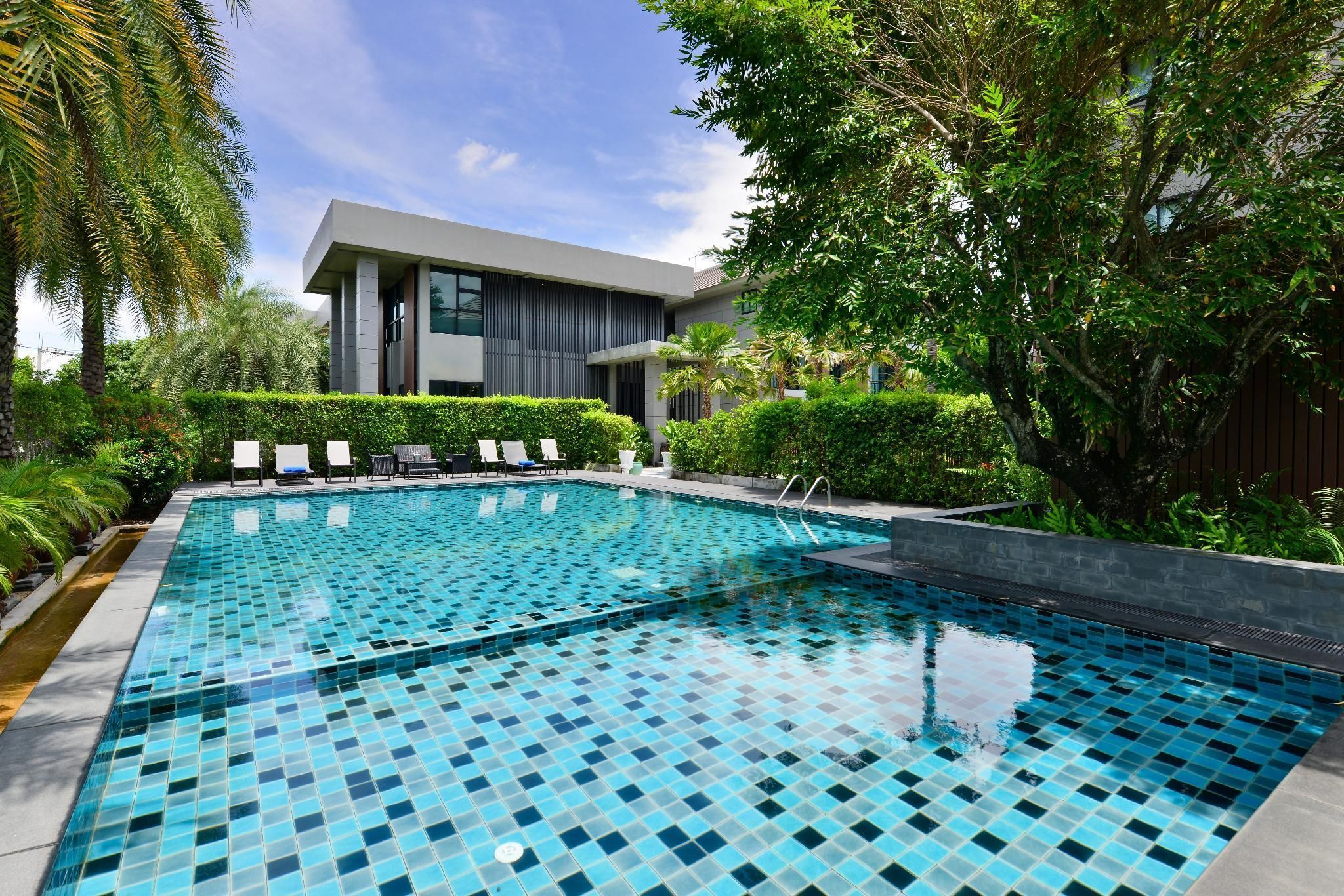 A modern swimming pool with a tiled surface, surrounded by lush greenery and a contemporary building in the background.