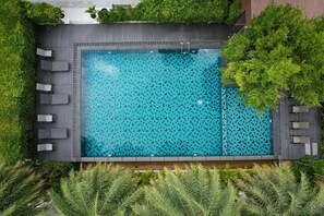 An aerial view of a rectangular swimming pool with a tiled mosaic pattern, surrounded by a wooden deck with seating and lush greenery. - Raweewan Residence (Bangkok)