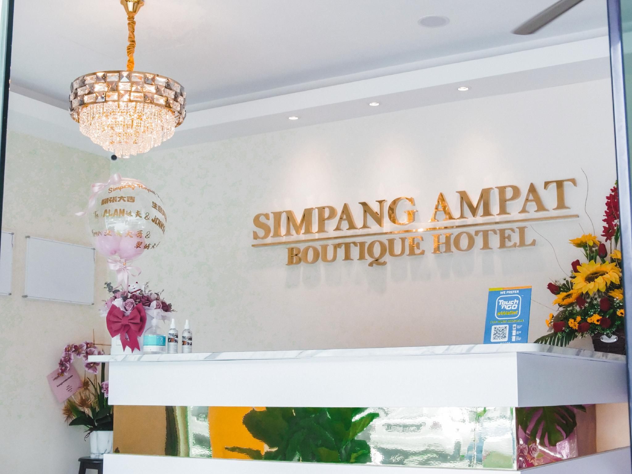 A reception area with a chandelier, a wall sign reading "SIMPANG AMPAT BOUTIQUE HOTEL", and a reception desk with a blue sign and a QR code.