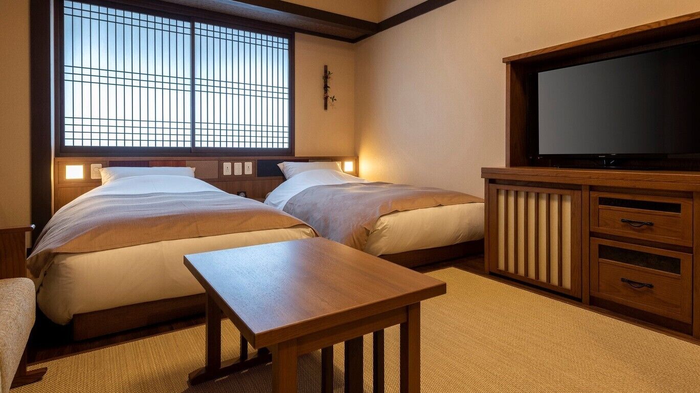 A room with two beds, a wooden table, a TV, and a large window with a grid pattern.