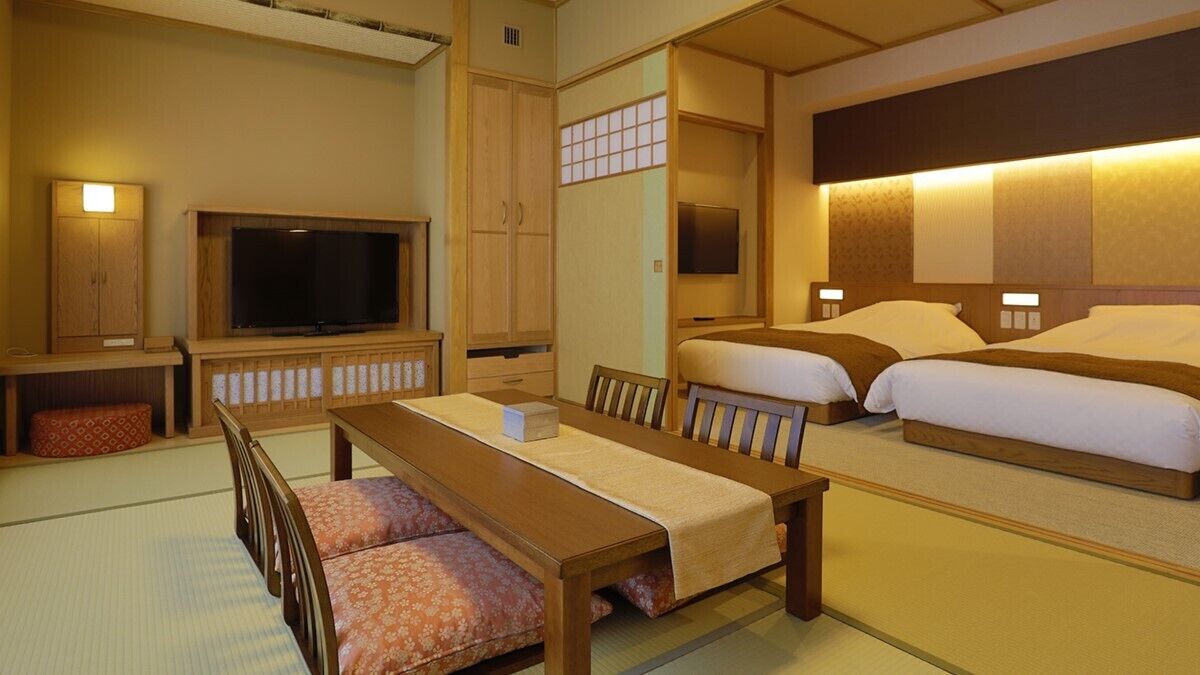 A traditional Japanese-style hotel room with tatami flooring, a low table, two beds, a television, and a sliding door.