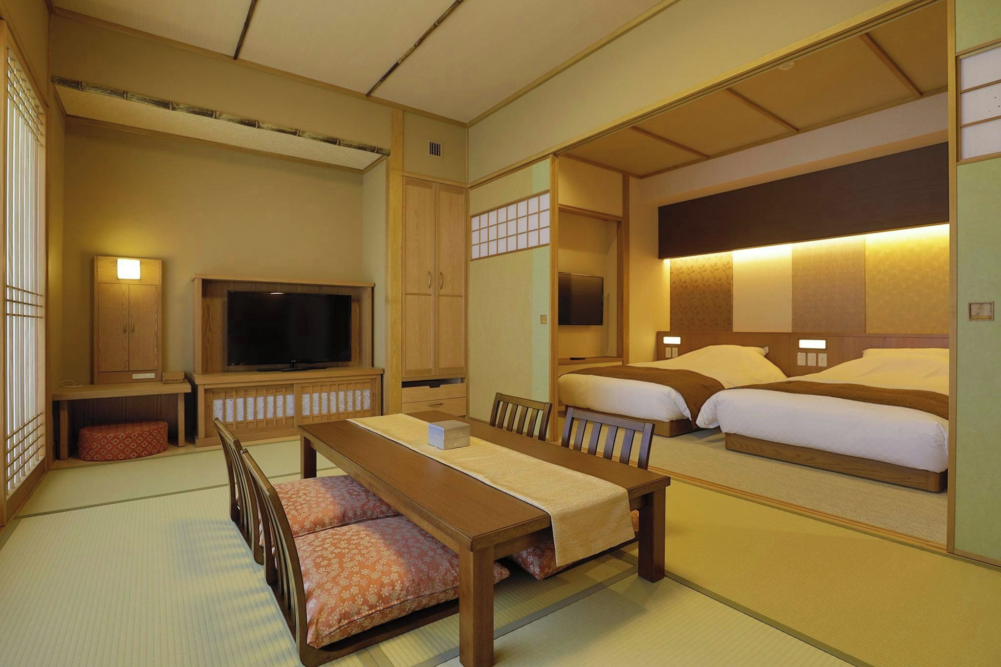 Una habitación tradicional japonesa con suelo de tatami, una mesa baja, dos camas con cabeceras, un televisor y una puerta corredera.